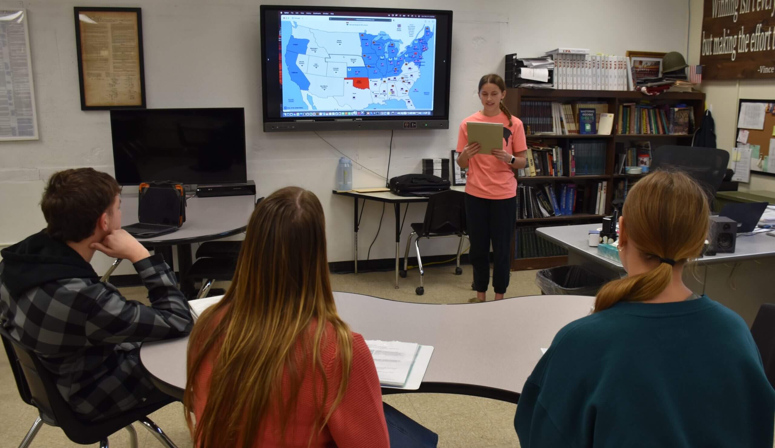 A high school student standing in front of a class, reading from a tablet and presenting a project featuring a digital map of the United States on a large screen.