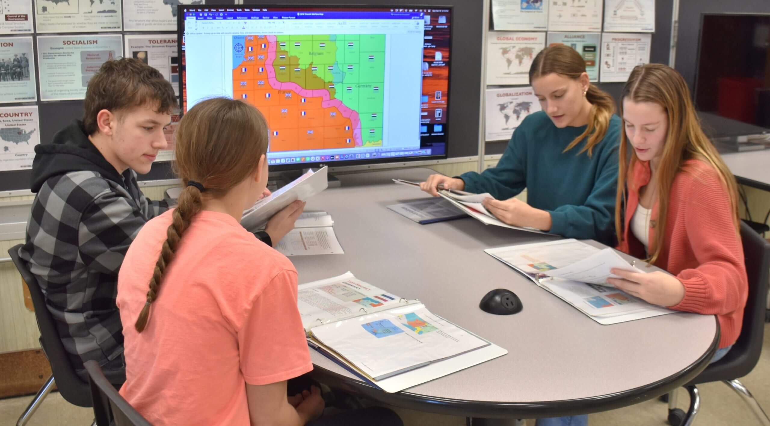 Four high school students sitting at a round table in a classroom, collaborating on a history project while looking at a digital map of Europe and individual binders