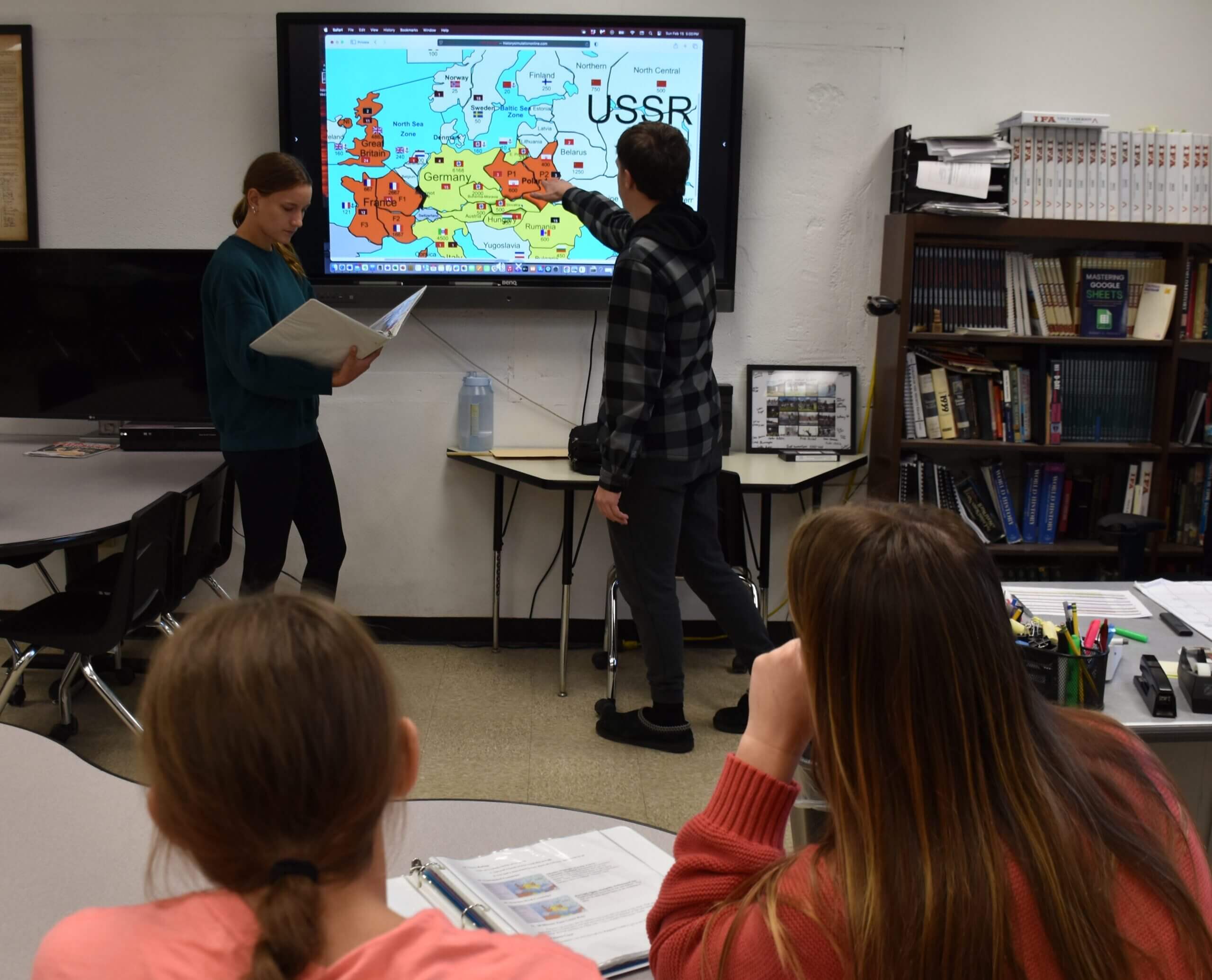 High school students studying world maps in hallway, pointing at Europe and Asia during geography activity